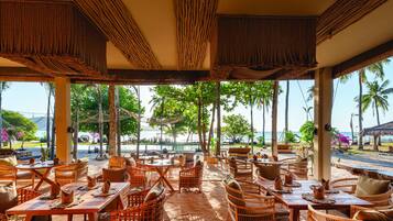 Un restaurante frente a la playa con mobiliario de mimbre, mesas con individuales y vistas al océano y palmeras.