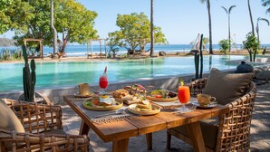 Un tavolo da pranzo in legno con cose per la colazione, un'area piscina con palme e vista sull'oceano.