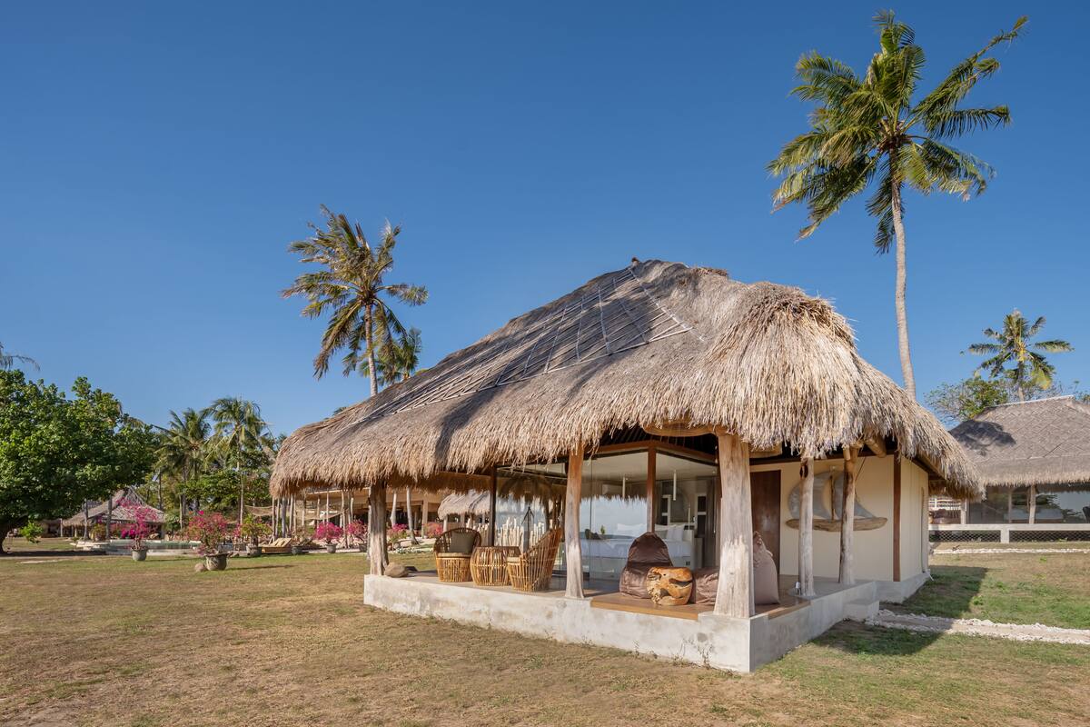 A thatched-roof hut with a porch, surrounded by palm trees and a clear blue sky.