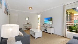 Ein modernes Wohnzimmer mit einem weißen Sofa, einem Couchtisch, einem Fernseher auf einem weißen Schrank und einem großen Fenster mit Blick auf ein Backsteingebäude.