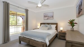 Ein Schlafzimmer mit Bett, Nachttisch, Schreibtisch, Deckenventilator, Fenster mit Vorhängen und einem Bild an der Wand.