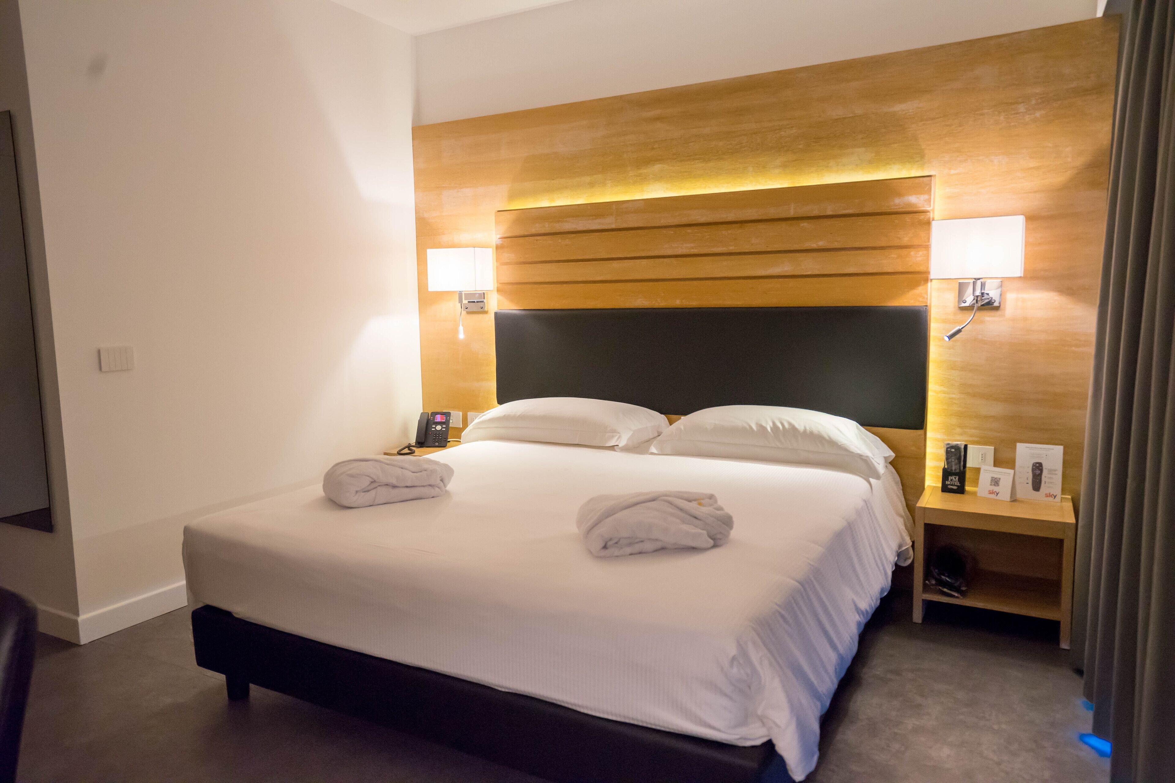 Une chambre d’hôtel avec un grand lit, deux lampes de chevet, une tête de lit en bois et une table de chevet avec un téléphone et un panneau.