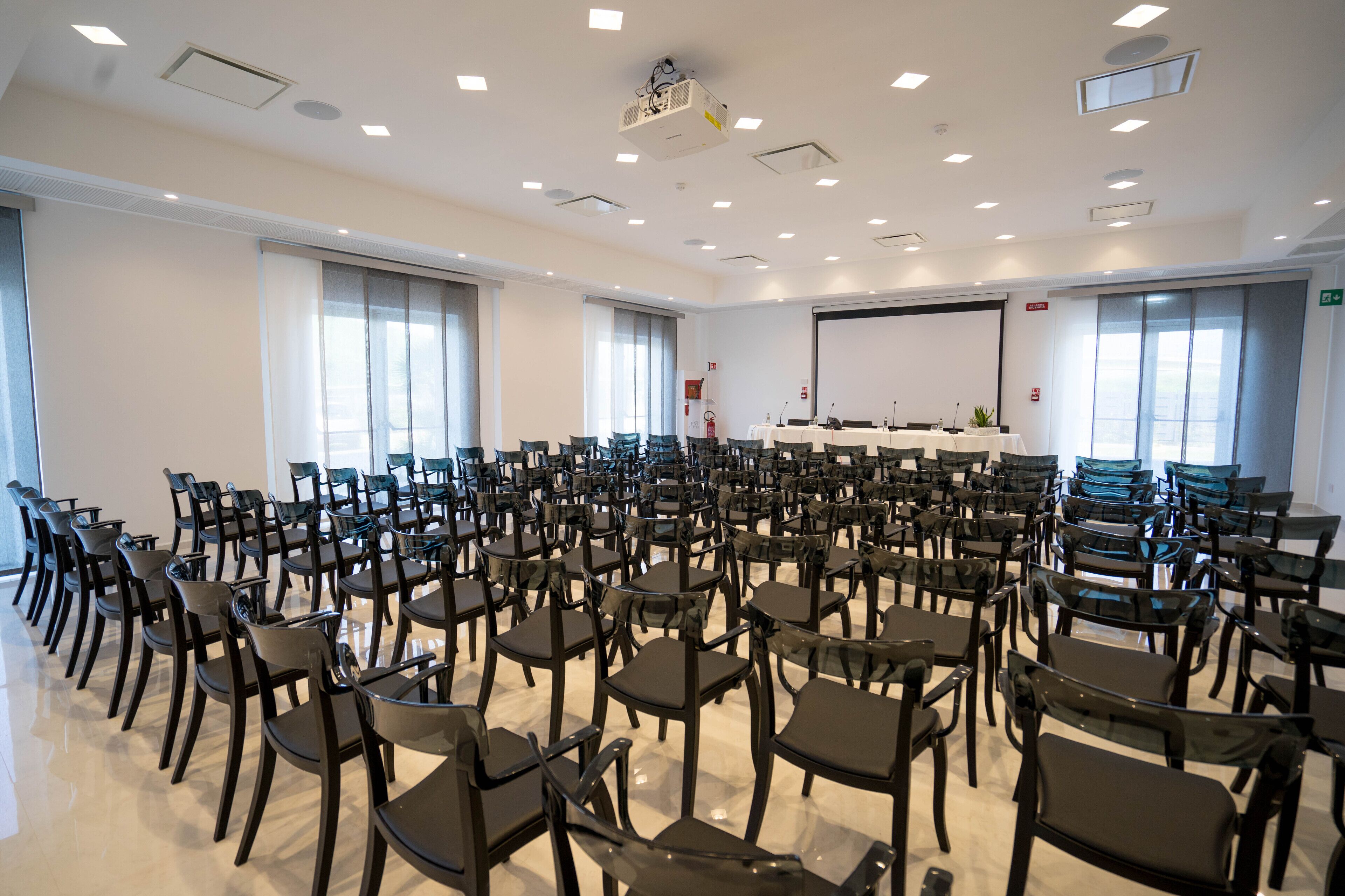 Un salón de conferencias con filas de sillas, una pantalla de proyector y un escenario con una mesa y sillas.