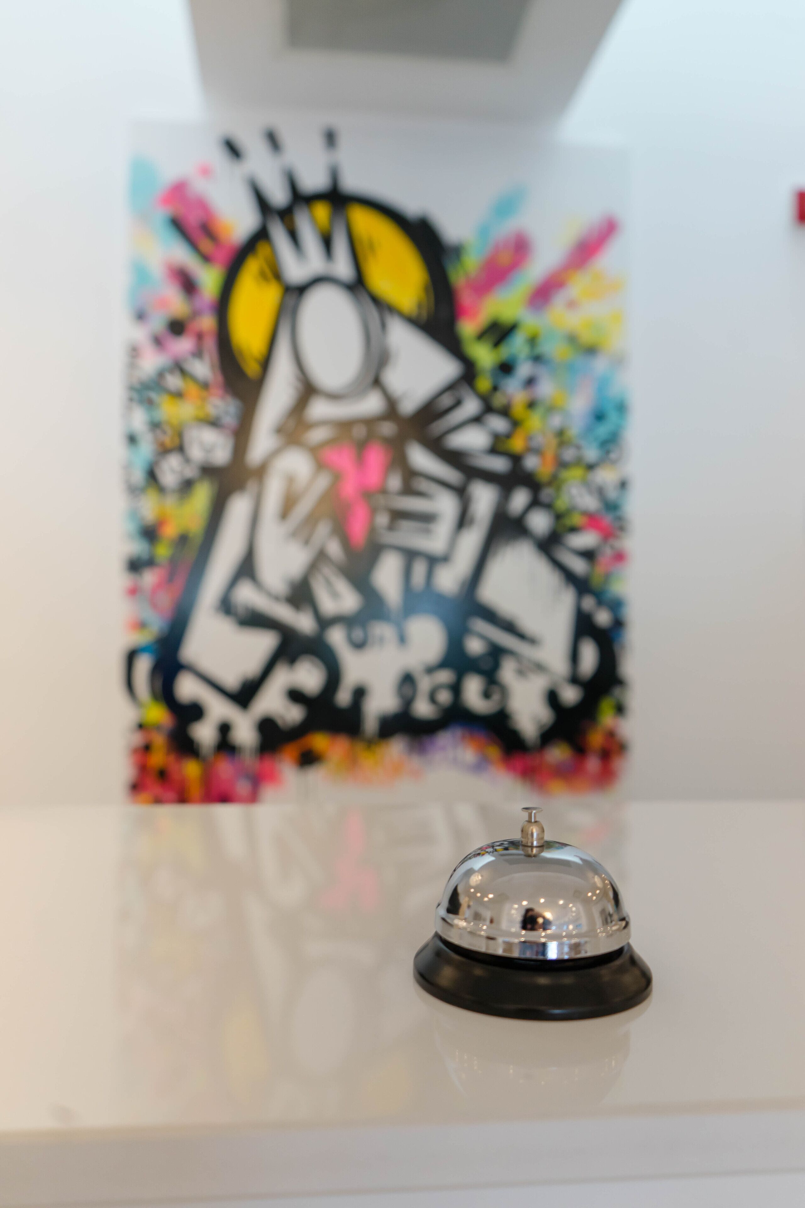 Un espace d’accueil d’hôtel agrémenté d’une œuvre de graffiti colorée sur le mur et d’une cloche argentée posée sur une surface réfléchissante.