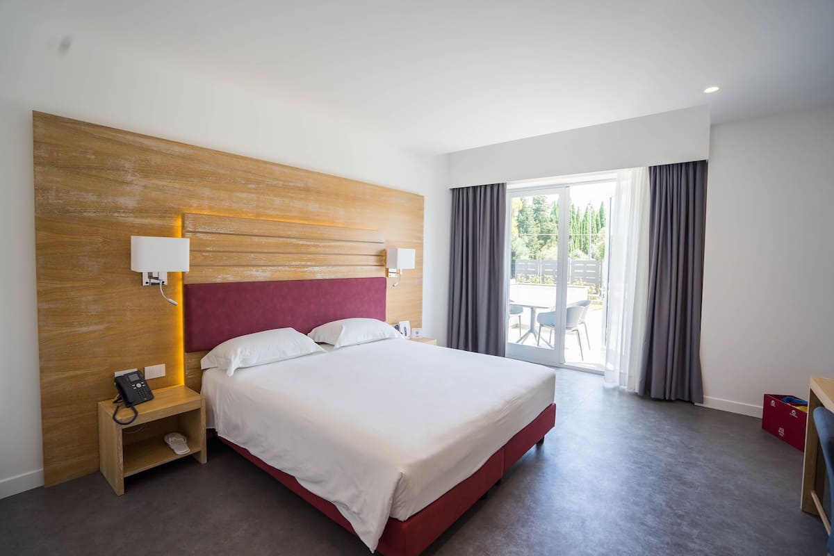 Ein Hotelzimmer mit einem großen Bett, einem Holzkopfschild, Nachttischlampen, einem Telefon auf einem Nachttisch und Blick ins Freie durch eine Schiebetür.