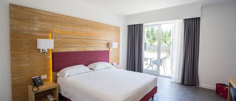 A hotel room with a large bed, wooden headboard, bedside lamps, a phone on a nightstand, and a view of the outdoors through a sliding glass door.