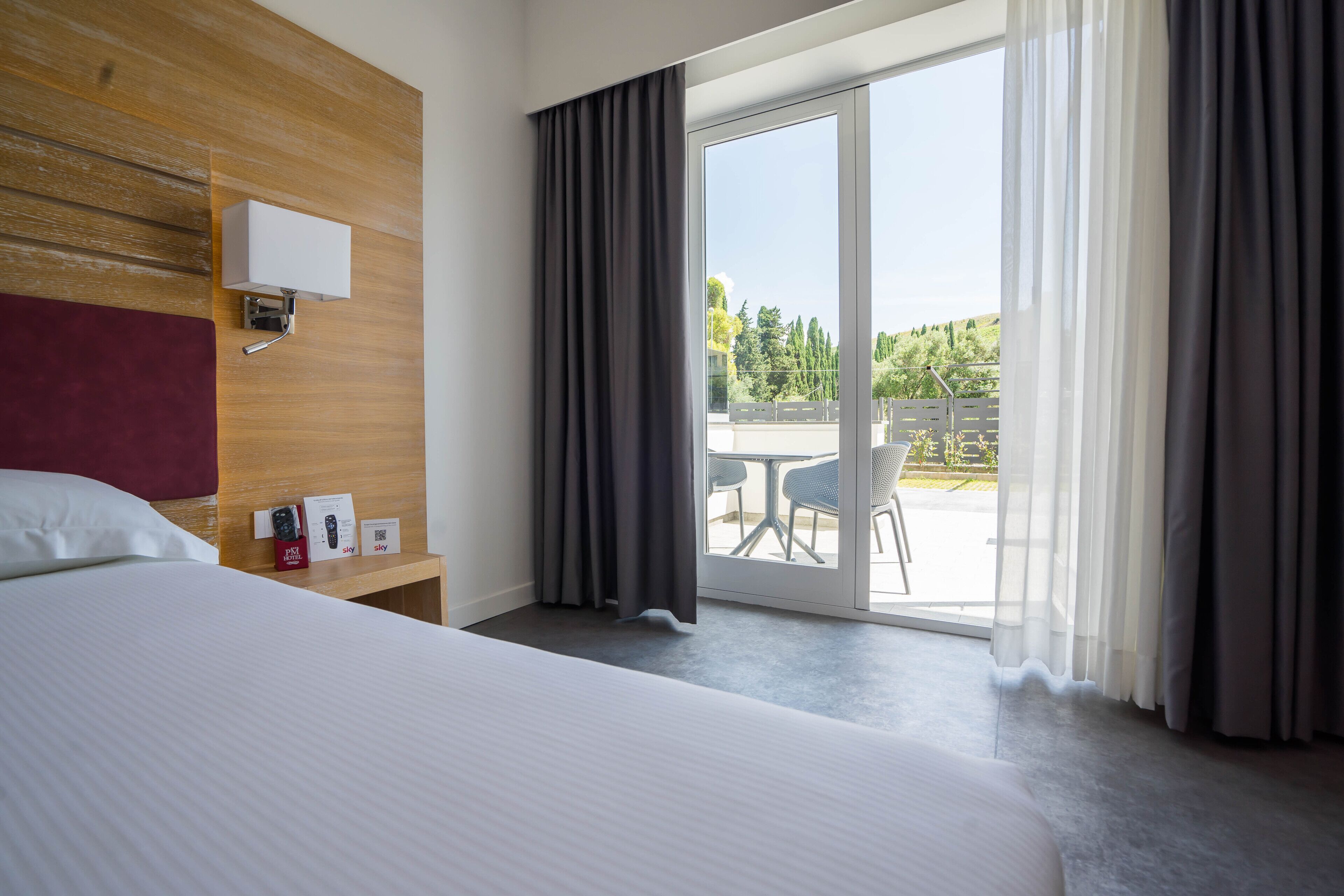 A hotel room with a large bed, a wooden headboard, a bedside table with a remote control, a brochure, and a view of the outdoor area through a sliding glass door.