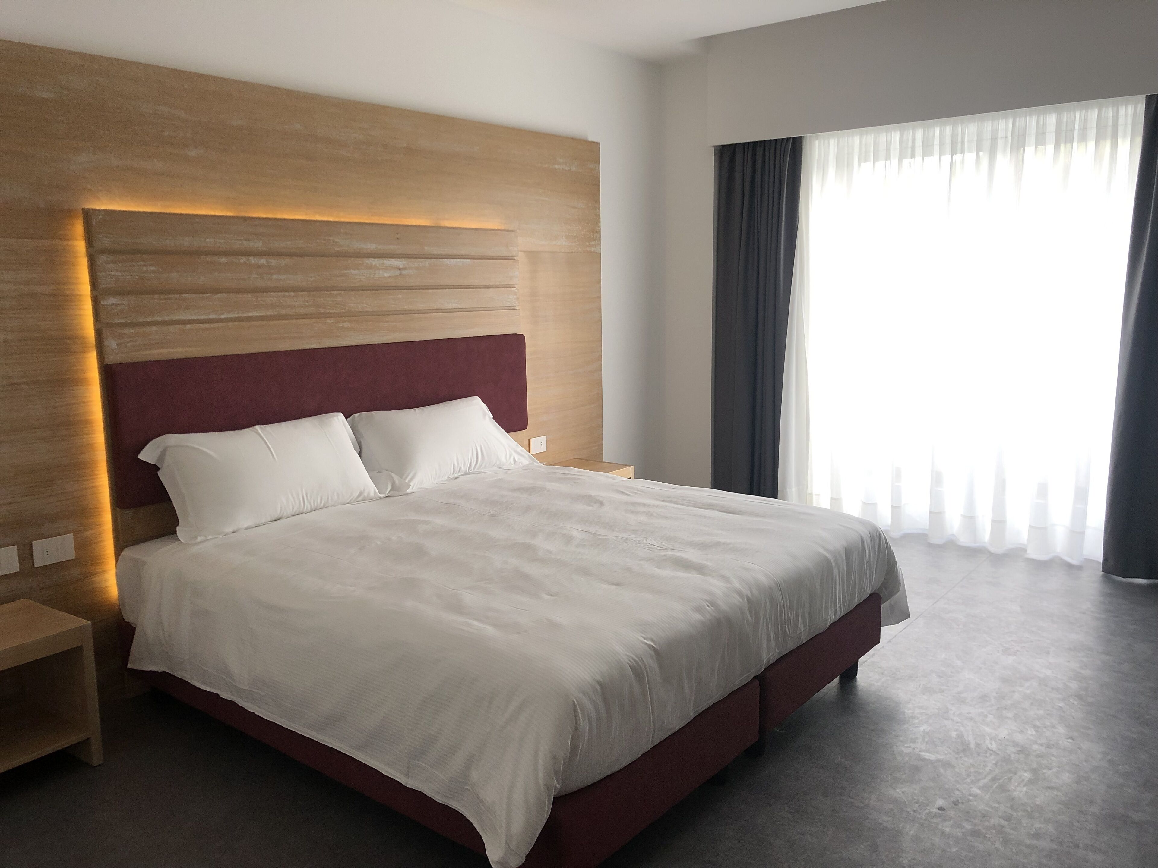 Una habitación de hotel moderna con una cama grande, cabecera de madera, mesita de noche y un ventanal con cortinas.