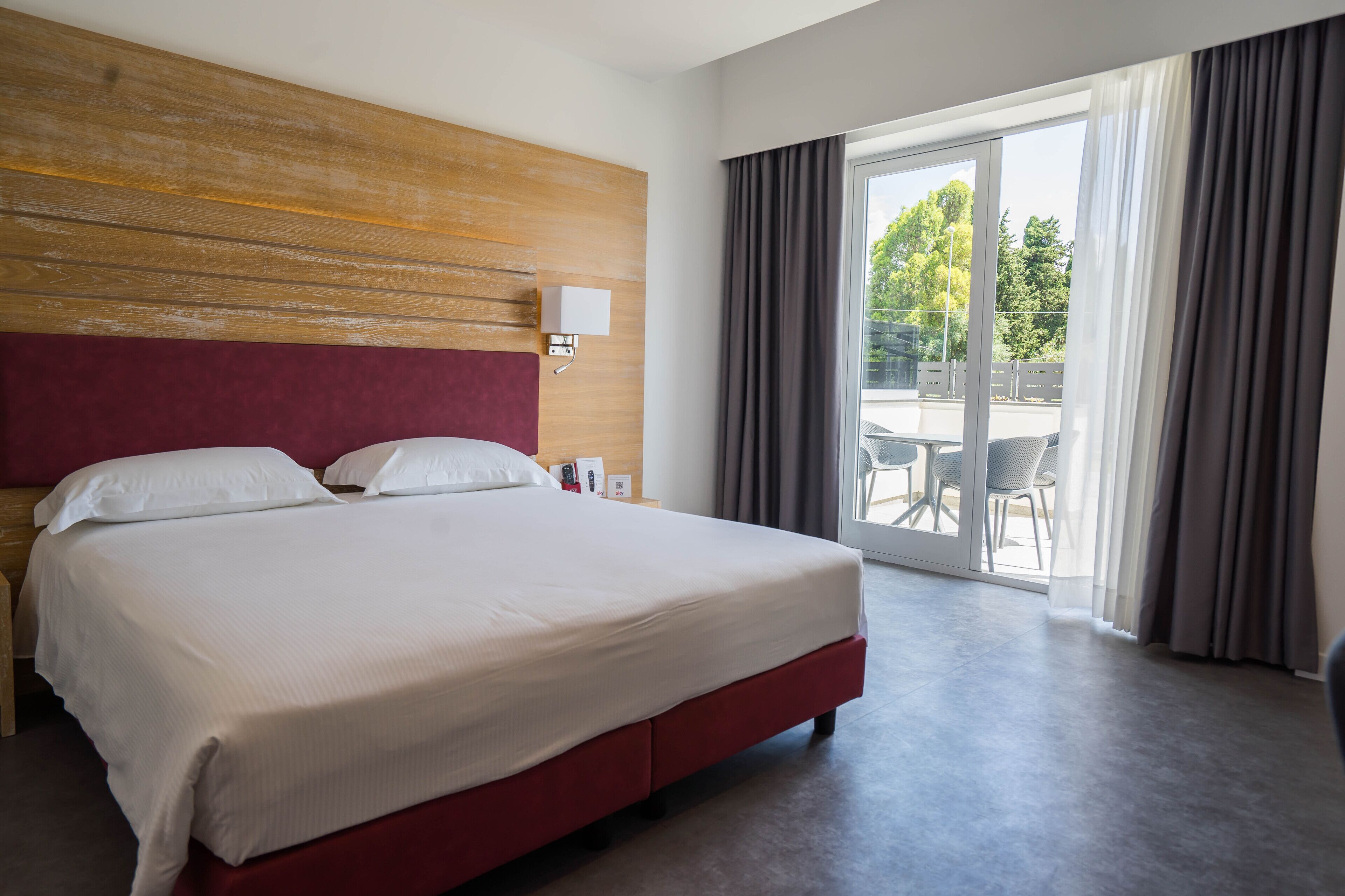 A hotel room with a large bed, a wooden headboard, a nightstand, a lamp, and a view of the outdoors through a sliding glass door.
