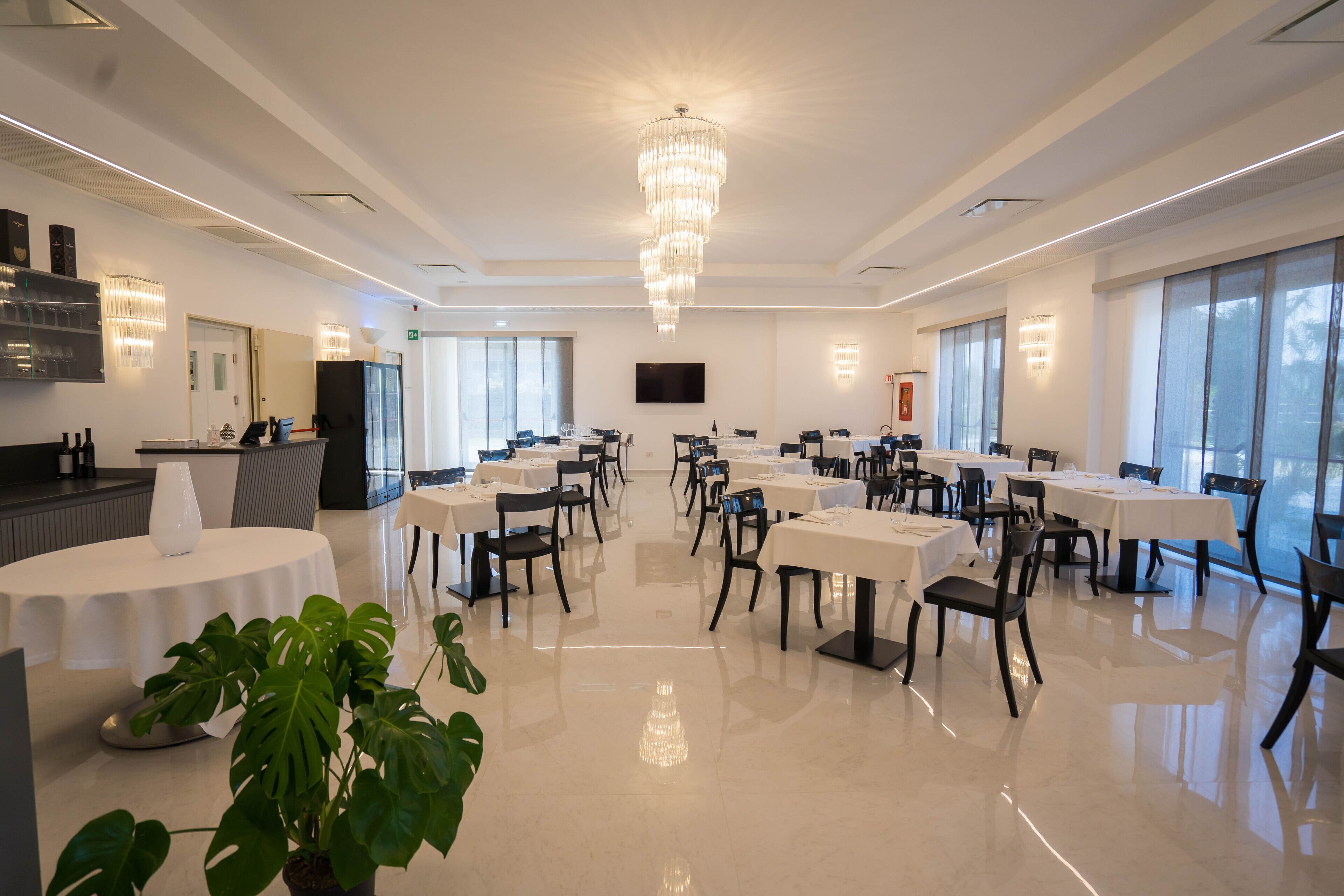 Un comedor amplio y luminoso con varias mesas y sillas, un gran candelabro y ventanas grandes que dejan entrar luz natural.