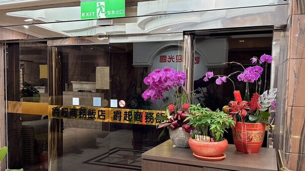 A hotel entrance with a green exit sign, a reception desk with potted plants, and a tiled floor. - Duke Business Hotel (Taoyuan City)