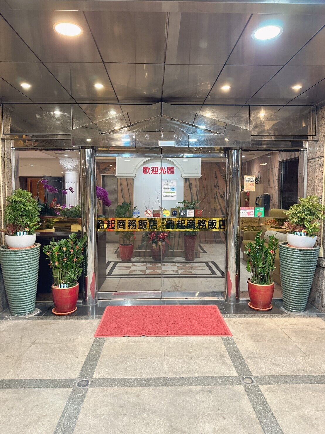 A hotel entrance with a glass door, potted plants, and a red carpet.