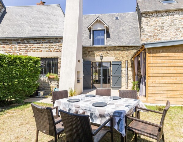 Une terrasse avec une table dressée pour quatre personnes, entourée de chaises en osier, devant un bâtiment en pierre doté d’une porte en bois et d’une fenêtre.