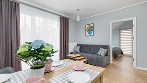 A modern living room with a grey sofa, a dining table set with pink and white plates, and a potted plant. - Climate Apartment | Balcony, Parking, 500 m to the Beach (Miedzywodzie)