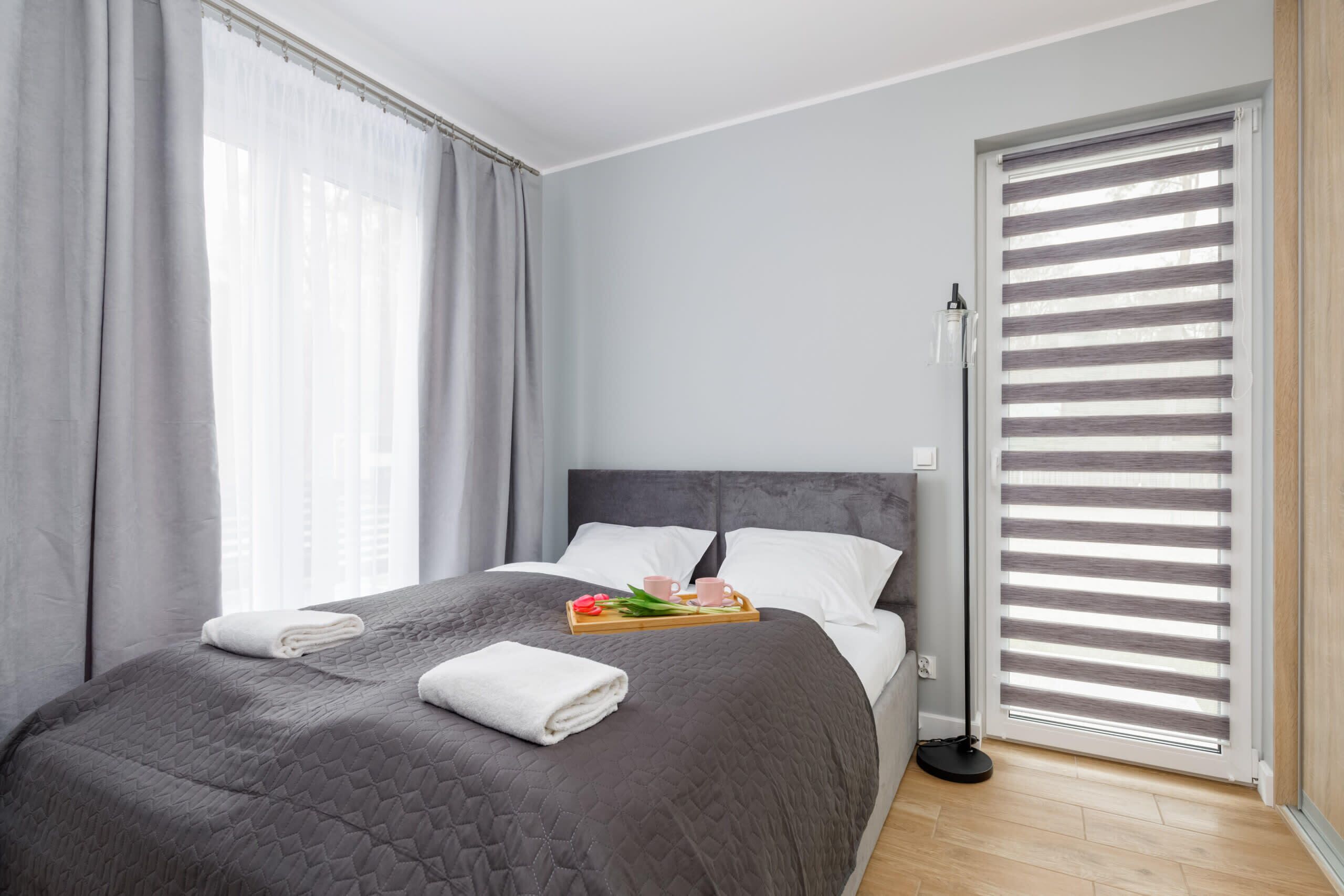Ein modernes Hotelzimmer mit einem großen Bett, grauen Vorhängen, einem Nachttisch mit Frühstücksgegenständen und einem Fenster mit Jalousien.