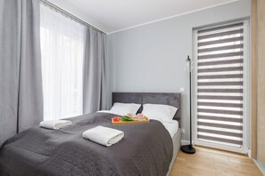 Une chambre d’hôtel moderne avec un grand lit, des rideaux gris, une table de chevet sur laquelle se trouve un plateau avec des articles pour le petit-déjeuner, et une fenêtre avec des stores.