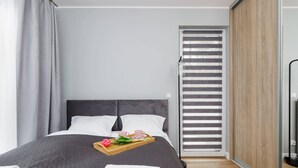 Een moderne slaapkamer met een groot bed, grijze gordijnen, een houten nachtkastje met een dienblad ontbijtartikelen en een schuifdeur van glas.