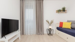 Una sala de estar moderna con un televisor de pantalla plana sobre un soporte blanco, un sofá con cojines de colores, una mesita auxiliar con una lámpara y una estantería con plantas en la pared.
