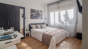 A modern bedroom with a bed, a dining table with plates and glasses, a dark wardrobe, a painting on the wall, and a window with sheer curtains.
