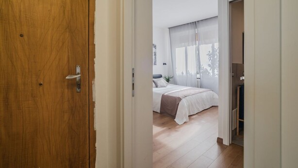 A bedroom with a bed, a window with sheer curtains, and a wooden floor.