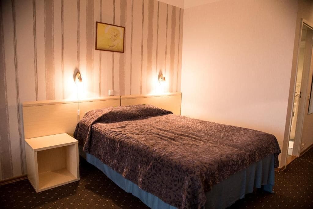 A hotel room with a bed, a nightstand, and a wall-mounted light fixture.