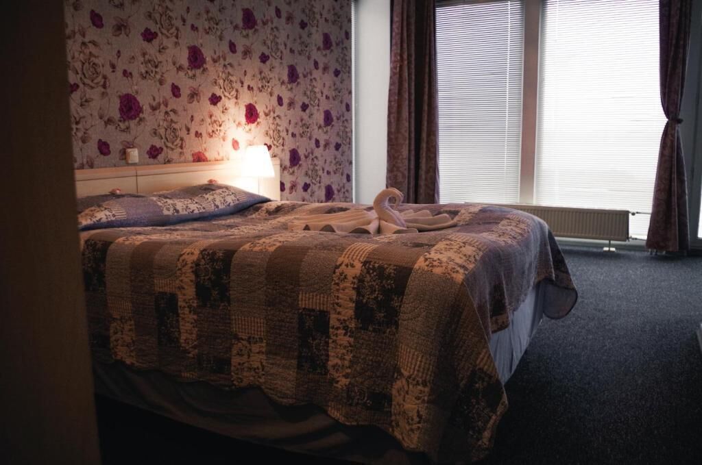 Ein ordentlich bezogenes Bett mit einer Steudecke, einer Nachttischlampe und einem floralen Wandpapier.