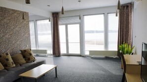 Una sala de estar moderna con un sofá gris, una mesa de centro de madera y un mueble para TV. Ventanas amplias con cortinas dejan entrar luz natural y ofrecen vistas al exterior.