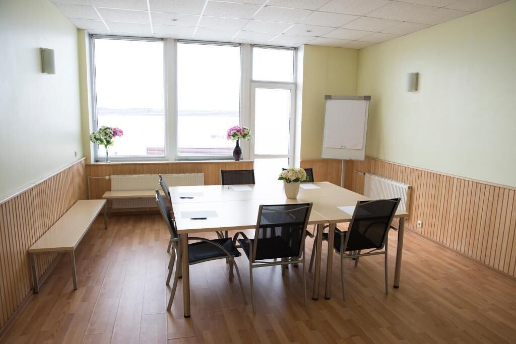 Un salón de conferencias con una mesa blanca, sillas negras, una pizarra y una ventana con flores.