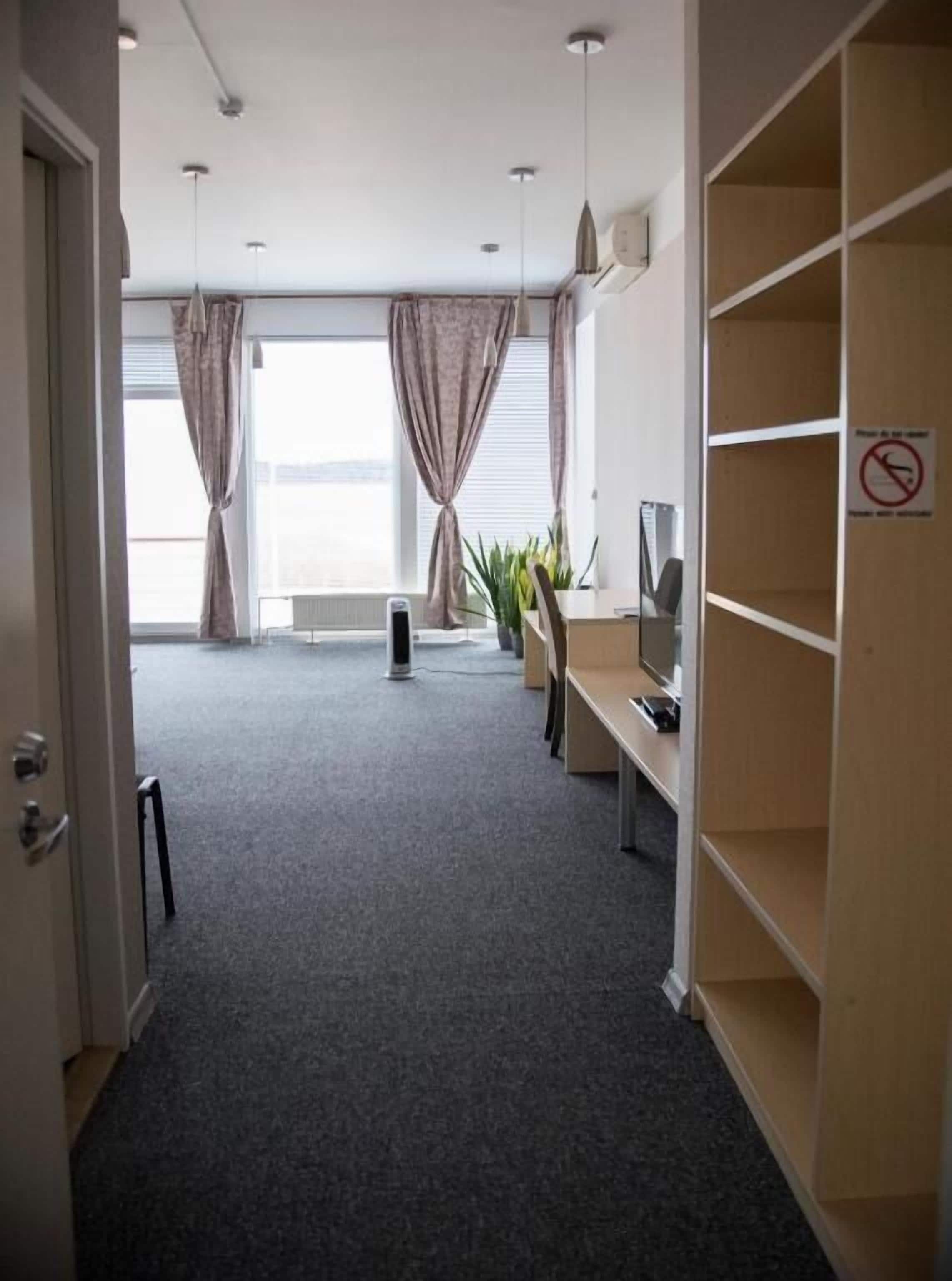 Une chambre d’hôtel avec un tapis gris, un bureau avec un ordinateur, une télévision et une étagère.