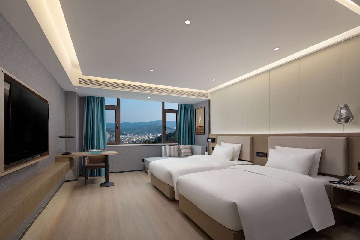 Une chambre d’hôtel moderne équipée de deux lits, d’un téléviseur à écran plat, d’un bureau avec une chaise et d’une grande fenêtre donnant sur la ville.