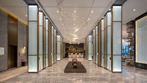 Un moderno vestíbulo de hotel con una escultura central, divisiones de vidrio y un mostrador de recepción al fondo.