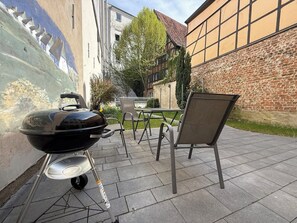 Een terras buiten met een barbecue, metalen stoelen en een tafel.