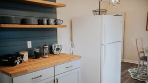 Una cocina con encimera de madera, armarios blancos y un refrigerador blanco. Hay estantes sobre la encimera con platos y tazas.