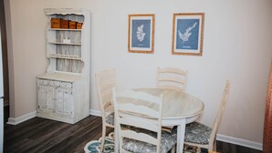 Un comedor con una mesa y sillas blancas, un mueble blanco con cajones y dos impresiones botánicas enmarcadas en la pared.