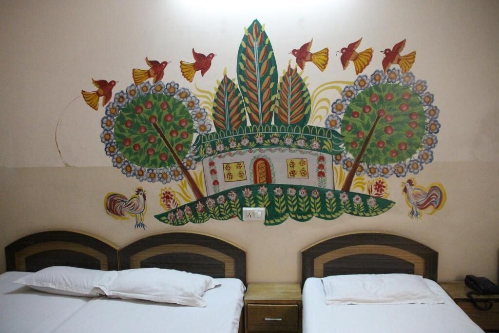 Une chambre d’hôtel avec deux lits simples, une petite table de chevet et une fresque murale représentant un arbre coloré, avec des oiseaux et un coq.