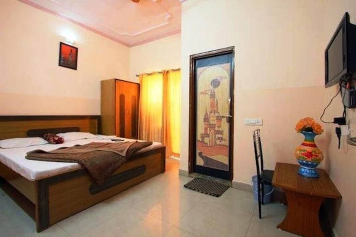 A hotel room with a bed, a desk with a vase, a chair, a television, and a door with a cityscape painting.