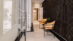 A modern hallway with a glass door, a seating area with a chair and a plant, and a dark patterned wall. - Urban Isle (Gzira)