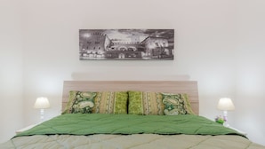 Ein Bett mit einer grünen Decke, zwei gemusterten Kissen, einem hölzernen Kopfteil und einem an der Wand hängenden Bild von einem Gebäude.
