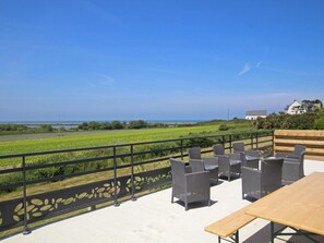 Een terras met rieten meubels en een houten tafel, met uitzicht op een groen veld en de zee.
