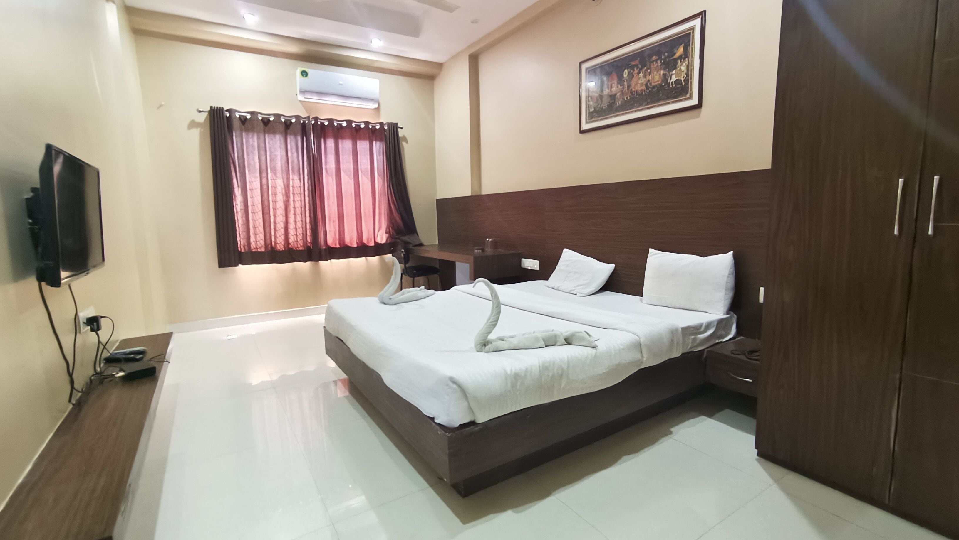 A hotel room with a bed, a desk, a chair, a TV, an air conditioner, and a painting on the wall.