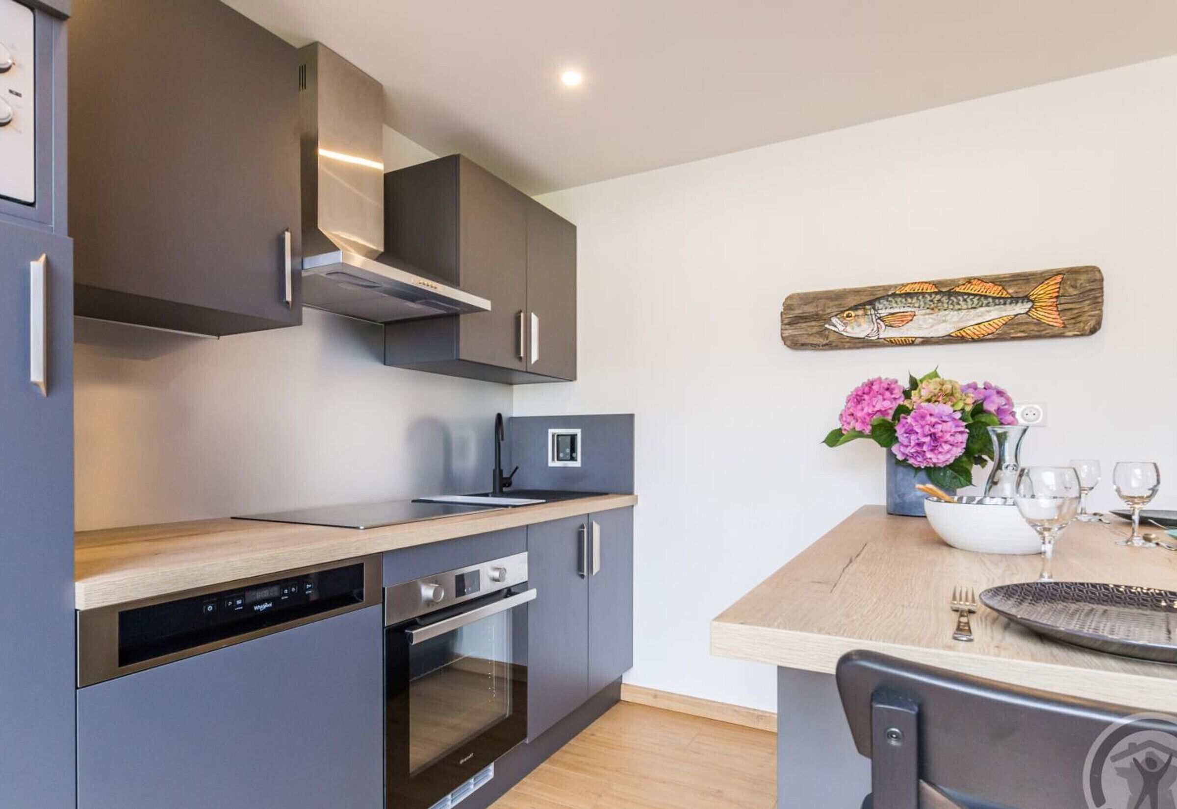 Une cuisine moderne avec des armoires grises, un plan de travail en bois et un four encastré. On y trouve également un coin repas avec une table en bois, des chaises et un vase de fleurs.