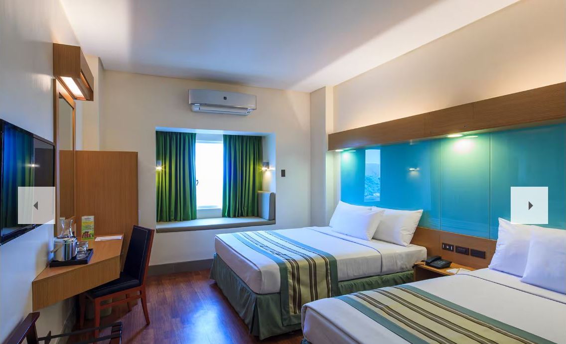 A hotel room with two beds, a desk, a chair, a window with curtains, an air conditioning unit, and a nightstand.