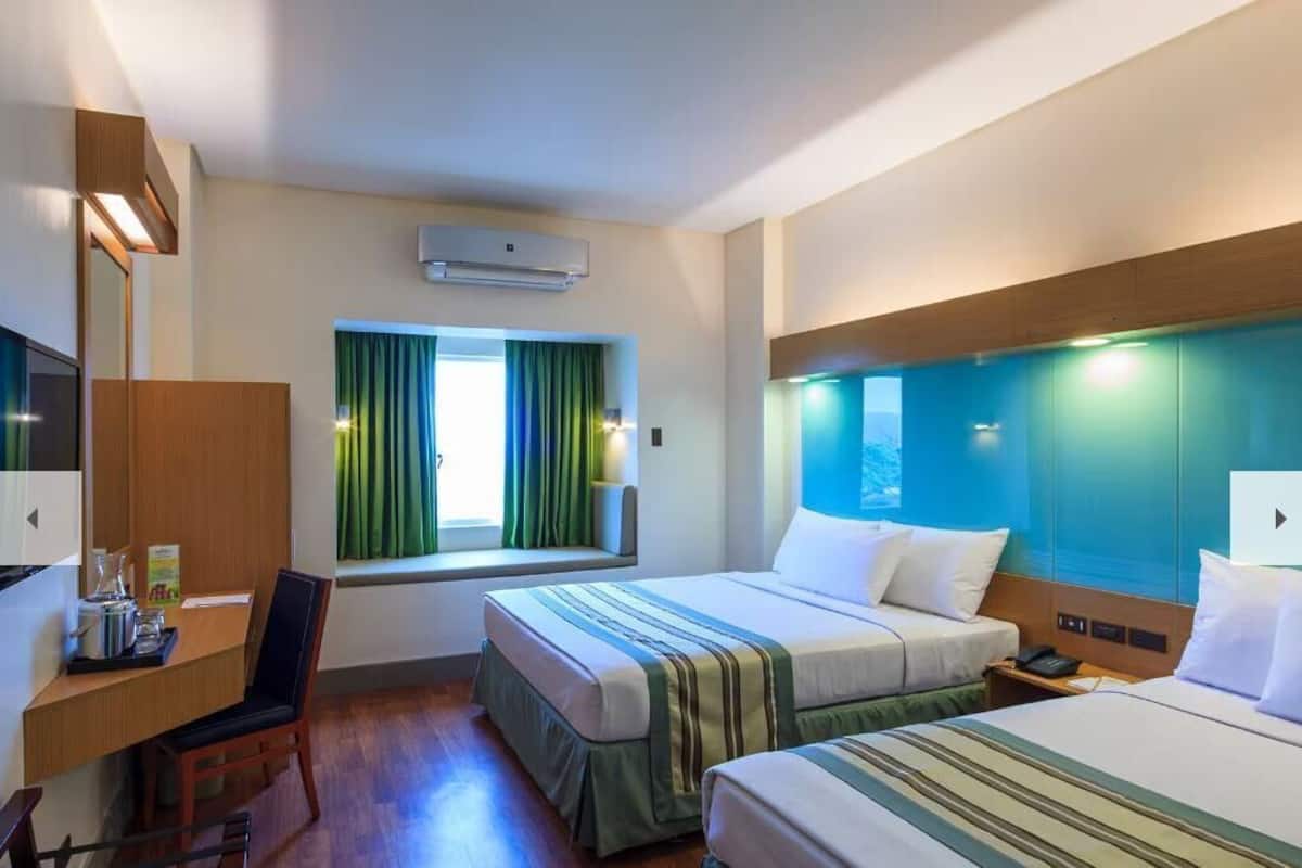 A hotel room with two beds, a desk, a chair, a window with curtains, an air conditioning unit, and a nightstand.