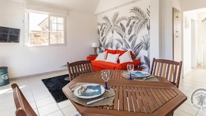 A dining area with a wooden table set for two, a red sofa with cushions, and a wall with a tropical leaf pattern.