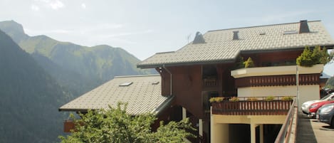 A chalet-style building with a sloped roof, wooden exterior, and a balcony with potted plants. It is situated in a mountainous area with a clear sky.