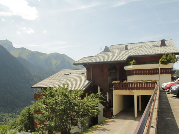 A chalet-style building with a sloped roof, wooden exterior, and a balcony with potted plants. It is situated in a mountainous area with a clear sky.