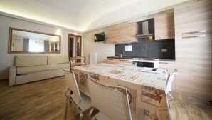 A compact kitchen with modern appliances, a dining table with a vintage tablecloth, and a sofa in a living area with a large mirror. - Tabor - STUDIO  sleeps 4 (Bardonecchia)