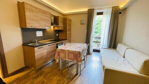 Una cocina funcional con armarios de madera, un fregadero y una mesa con mantel a cuadros. Un sofá se encuentra cerca de una ventana con cortinas.
