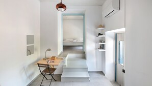 A narrow hallway with a wooden desk and chair, a hanging pendant light, a staircase leading to a bedroom, and a door with a blue frame.