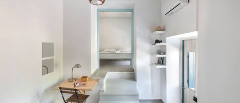 A narrow hallway with a wooden desk and chair, a hanging pendant light, a staircase leading to a bedroom, and a door with a blue frame.